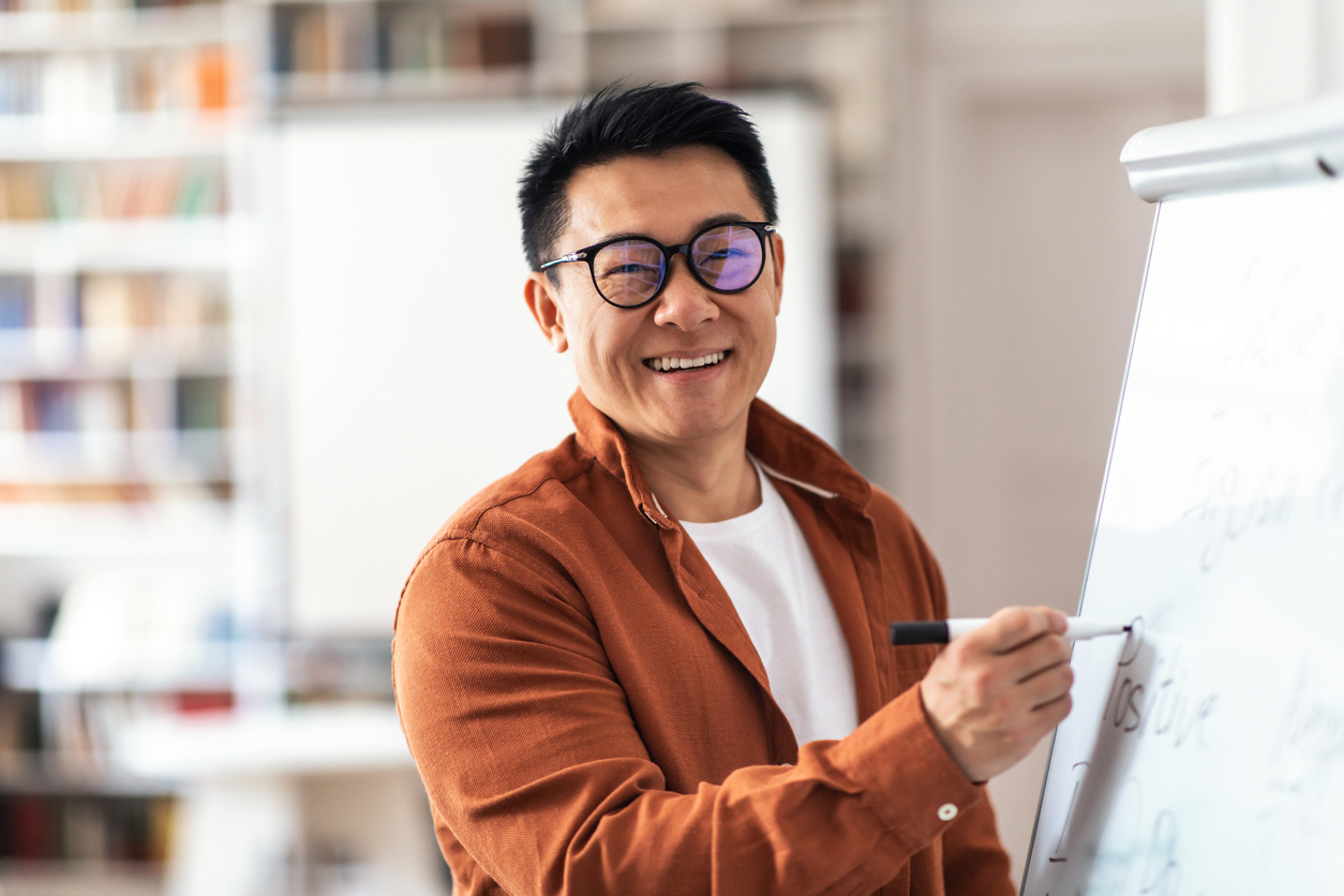Asian man with glasses smiling, writing on newsprint looking at viewer 