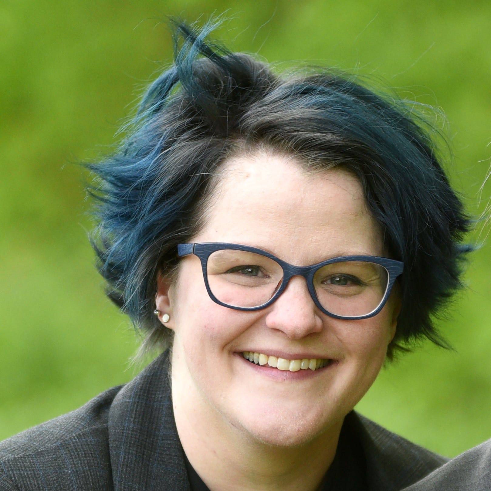 Emily Nagoski has blue and black hair, and wears stylish dark-rimmed glasses. She is smiling into the camera and the background behind her is blurred greenery.