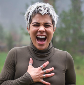 Lauren Hanner laughing into the camera with her hand on her heart, wearing a brown turtleneck