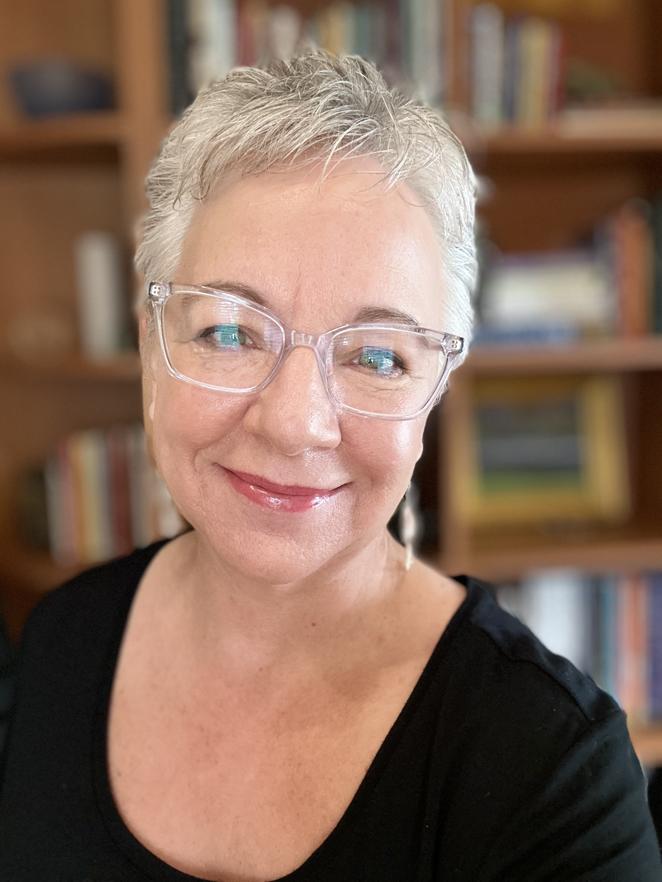 Melanie Davis has short, stylish white hair, clear glasses, and is smiling into the camera.