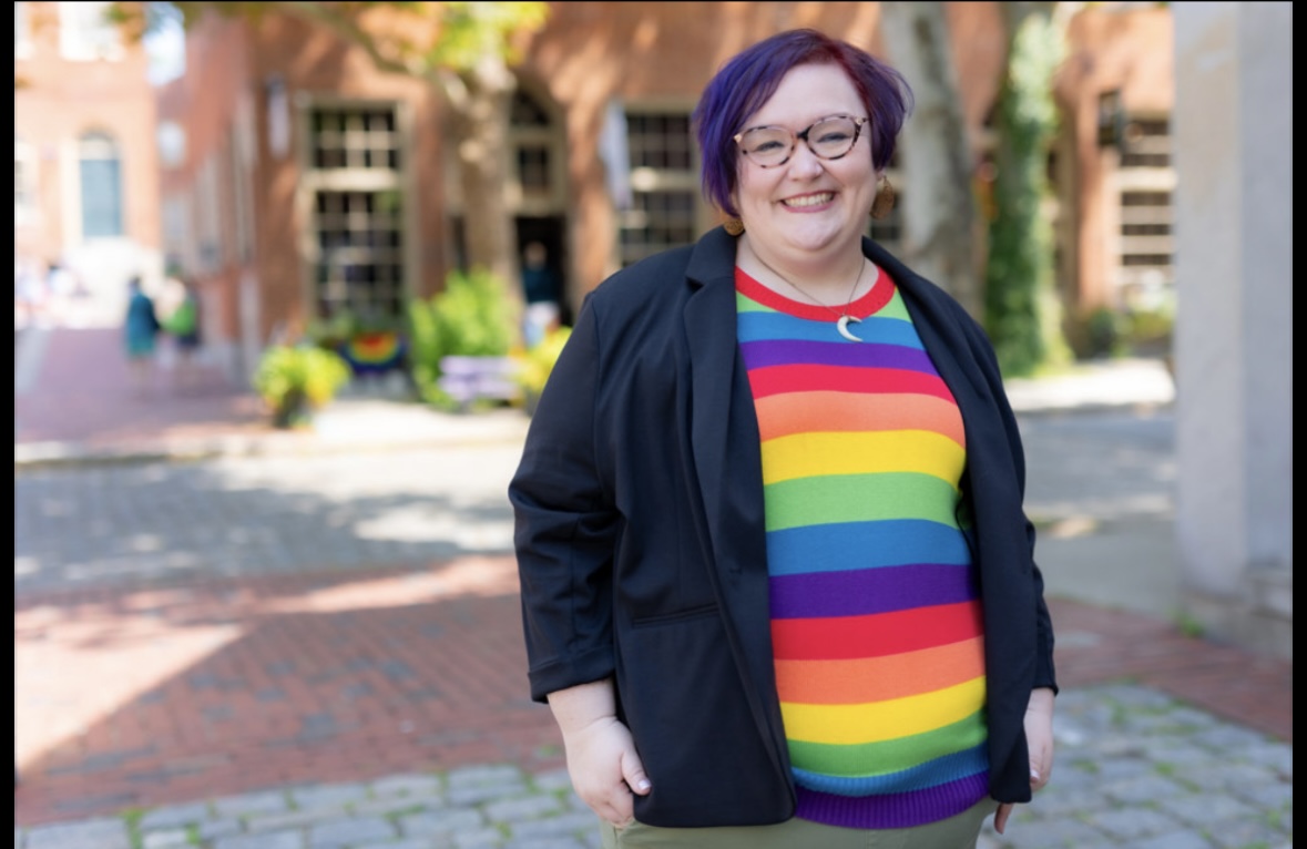 Miranda is standing on a street with a brick building behind her. She is wearing a rainbow colored striped shirt, has short dark hair and glasses.