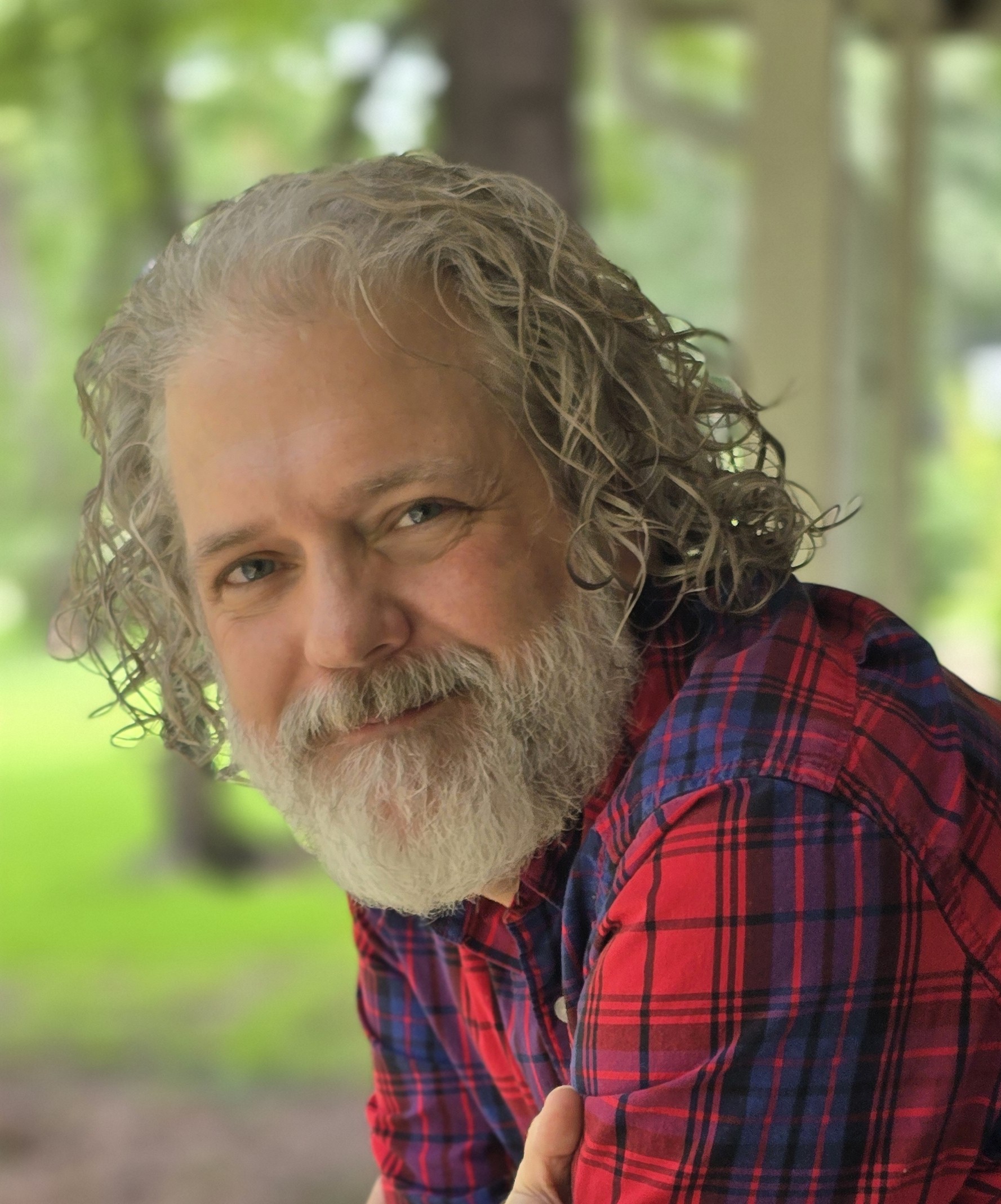 Marc Prevette Marc is leaning into the camera and smiling. He has sholder length gray hair and a thick gray beard. He is wearing a red plaid shirt.