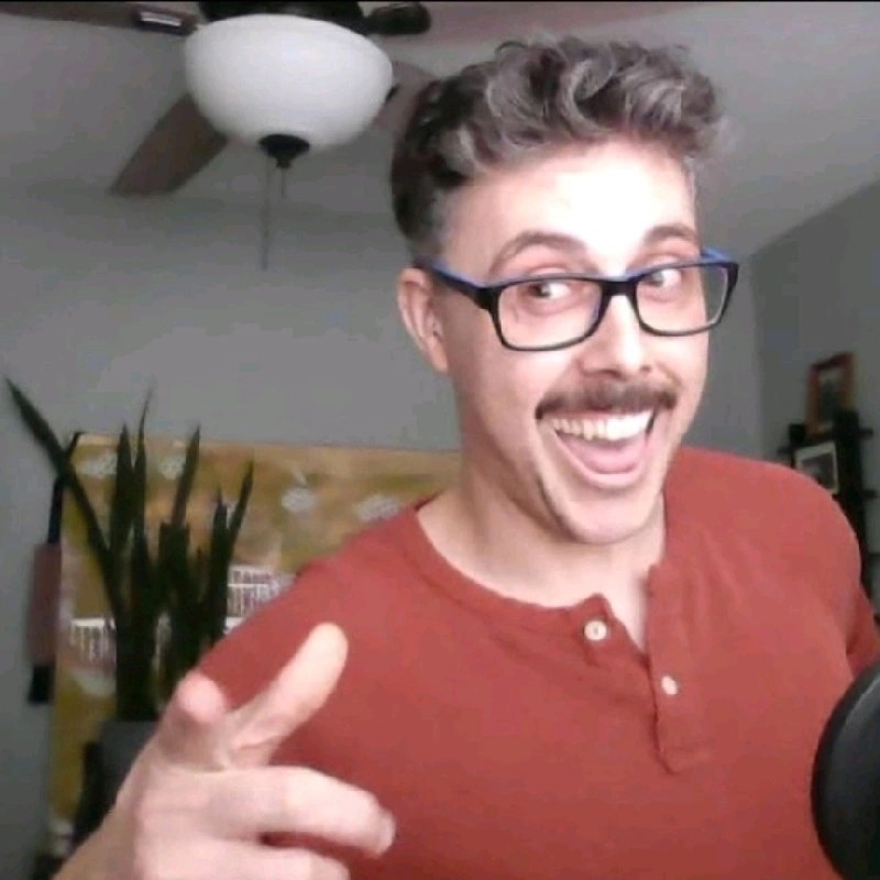 Photo of Nicholas with a big smile, pointing at the camera. He is wearing black stylish glasses and a red button down shirt