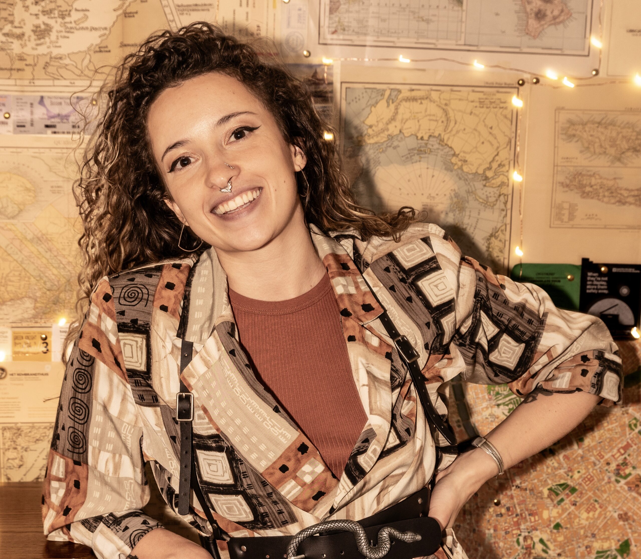 Vivi Mage has long, curly brown hair, brown eyes, and nose jewelry. She is leaning with one arm on a table and the other on her hip, smiling and looking up.