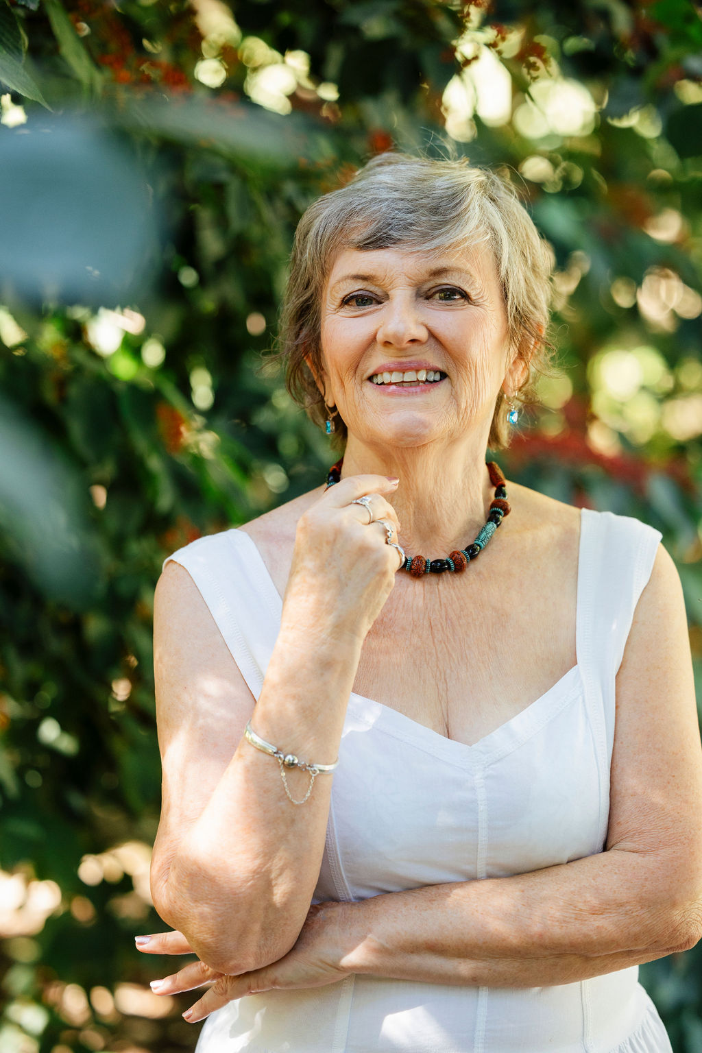 Rev Beverly Dale is standing in front of some greenery wearing a white summer dress, a colorful necklace and smiling. She is gray haired and green eyed.