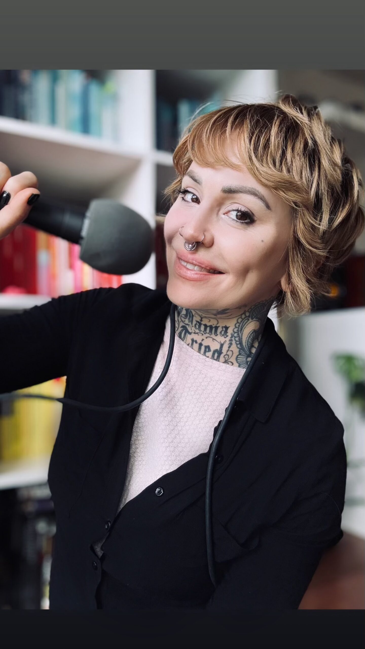 Elle Stanger has short wavy blonde hair, dark eyebrows, and dark brown eyes. She has a large tattooo that says Mama Tried on her neck. She has several nose piercings and a septum piercing. She is sitting in front of a bookcase in the background, smiling and holding a microphone.